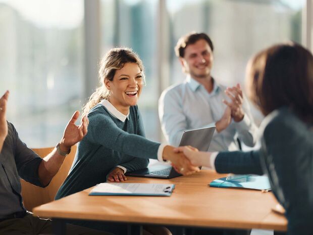 Two women shake hands joyfully across a table, a man claps in the background