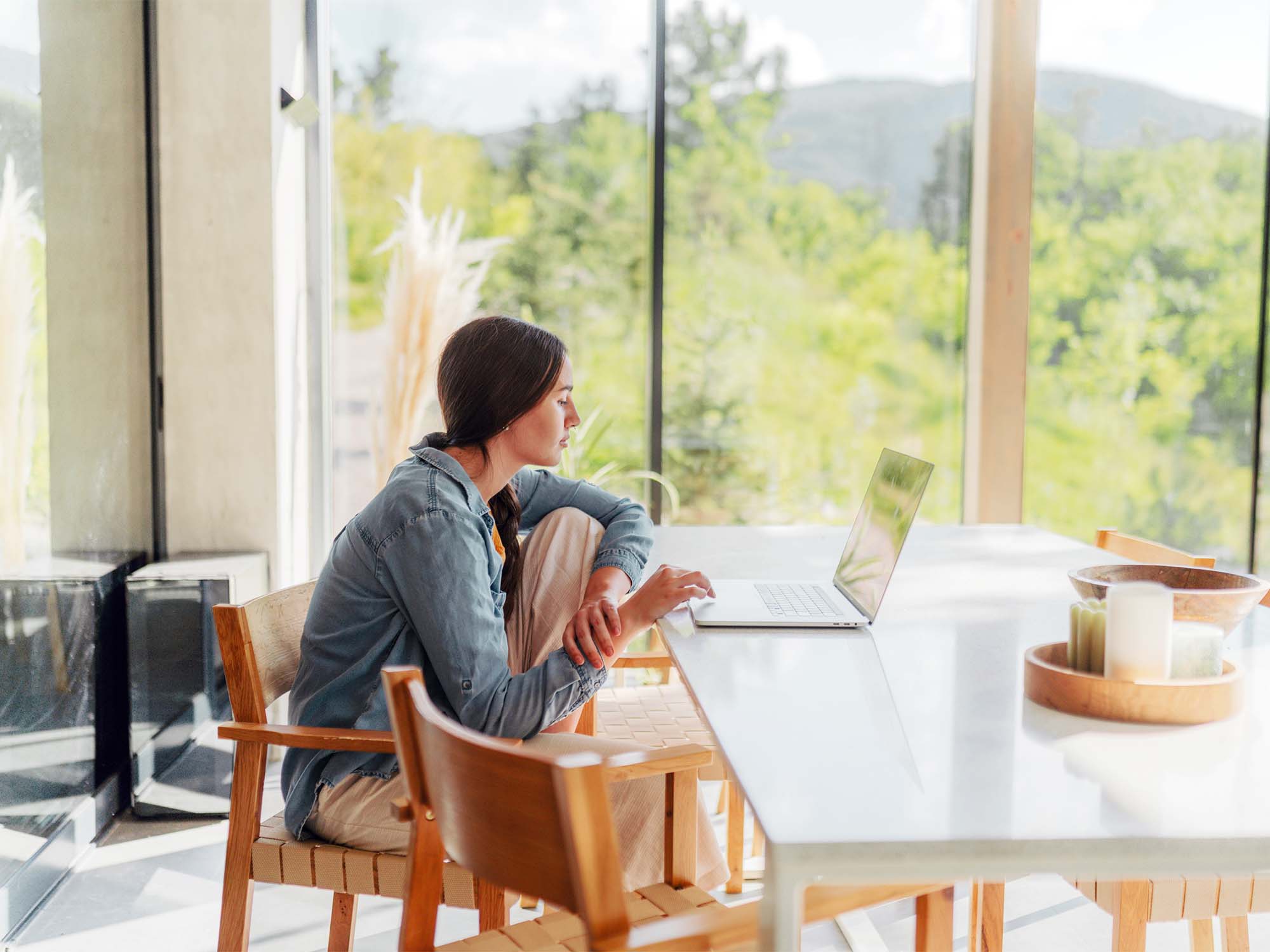 Woman sitting at a table with a laptop, behind her is a window with a view of the forest and mountains